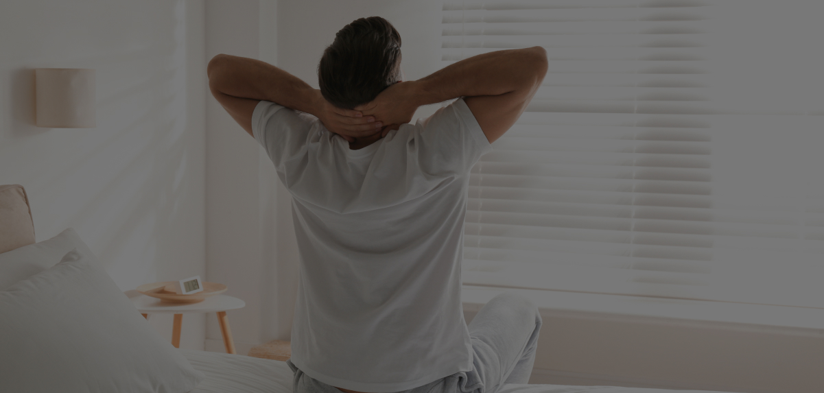 A man is sitting on his bed stretching in front of the window